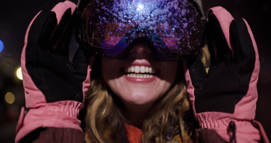 Close-up of a happy snowboarder on a snowy night at a ski resort. She smiles and looks at the camera while taking off her goggles.