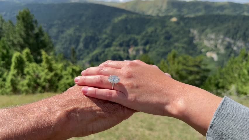 Landscape video of a couple gently holding hands in Montenegro highlighting a beautiful oval diamond engagement ring with mountains and greenery behind them.
