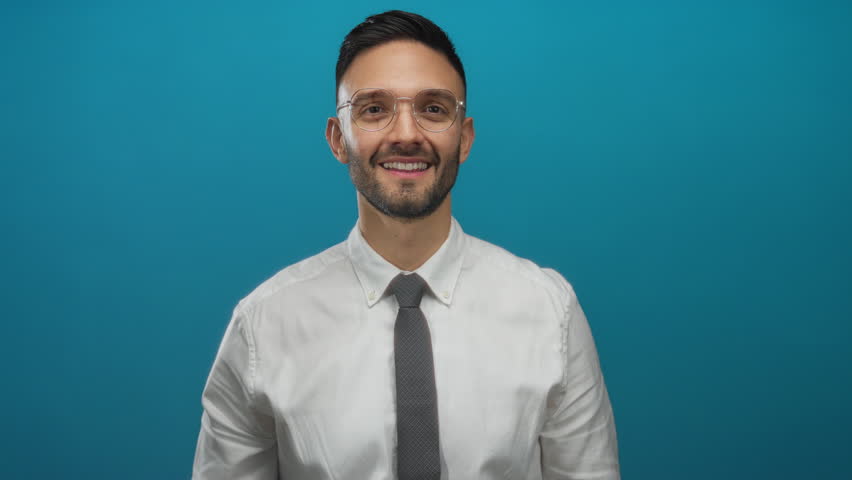 Young man with glasses and beard wearing white shirt and tie crossing fingers against blue background, expressing hope and expectation.