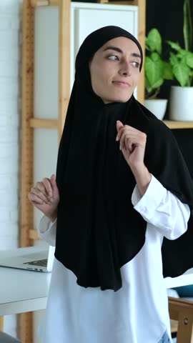 Vertical screen young muslim woman in black hijab dancing joyfully in a modern home office, smiling and celebrating freedom and happiness