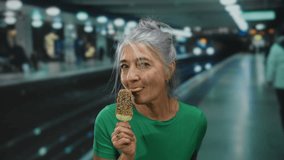 Senior woman with grey hair enjoying ice cream at an indoor train station, capturing a joyful moment amidst urban transit with a vibrant background. - Powered by Shutterstock - Get 15% off with code: PIKWIZARD15