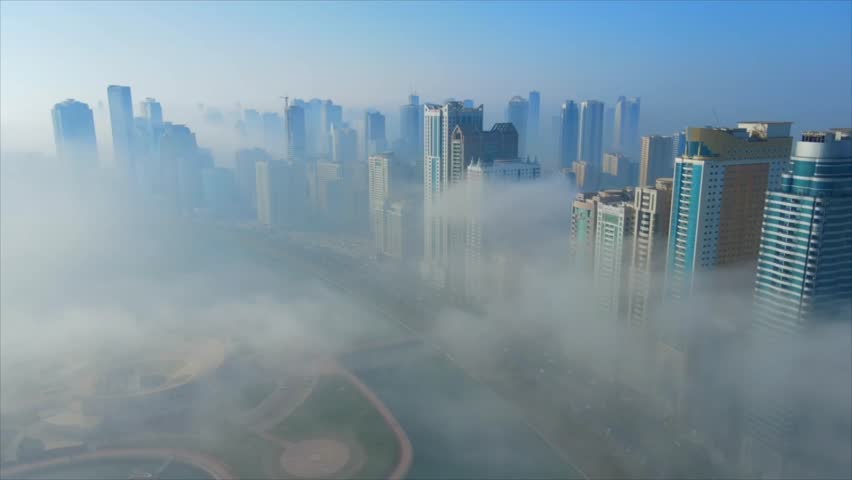 A dramatic aerial shot captures the modern skyscrapers of Sharjah, UAE, as they pierce through a dense layer of low-lying morning fog. Sharjah, UAE, 14 March 2025