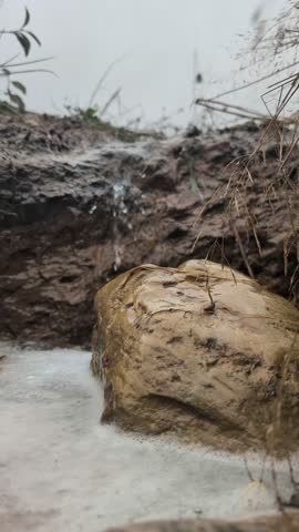 Close-up of a small natural stream flowing over wet rocks and soil after rainfall, capturing raw earth textures, foam, and gentle water movement in overcast light.