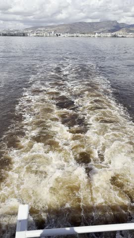 Wake trail of a moving boat on the sea with city skyline and mountains on the horizon under cloudy sky. High quality 4k footage
