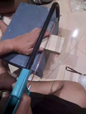 A person saws a small piece of light-colored wood, using a hacksaw or mini handsaw propped against a dark block of wood on a tiled floor.