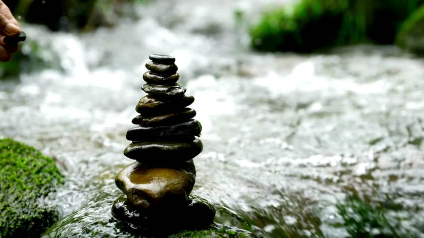 A hand carefully places the final stone on a balanced stack in a flowing river. A zen concept of stability, patience, mindfulness, and harmony.