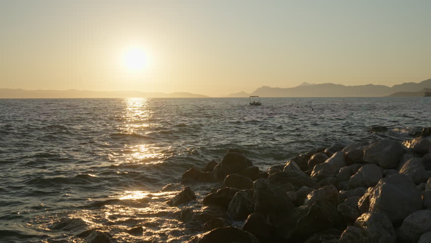 The golden sun sets over the Adriatic sea, creating a beautiful reflection on the water. Waves splash on a rocky shore in Croatia, with a small boat visible on the horizon. A romantic summer scene.