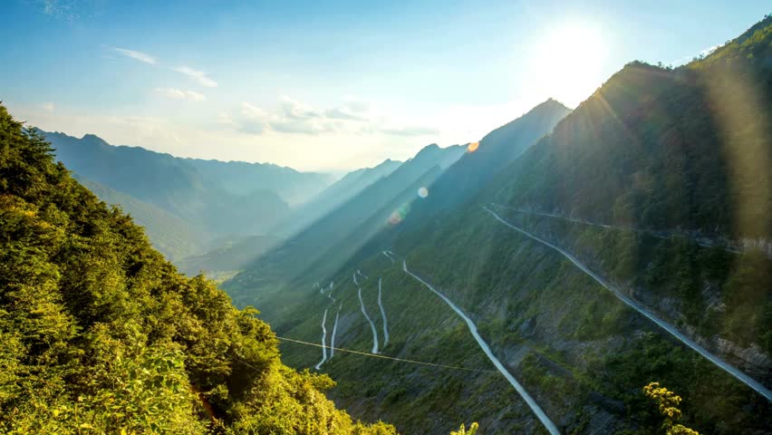 A stunning timelapse of the sun setting over a majestic mountain range. A long, winding road with switchbacks descends into the green valley below.