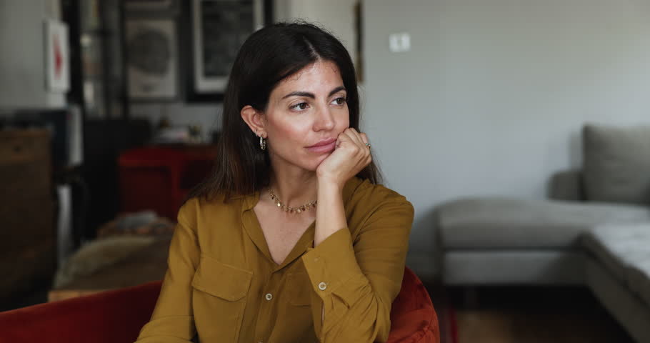 Bored pensive young Latin homeowner woman looking away, resting in armchair, touching chin, thinking, daydreaming, suffering from procrastination, frustration, boredom