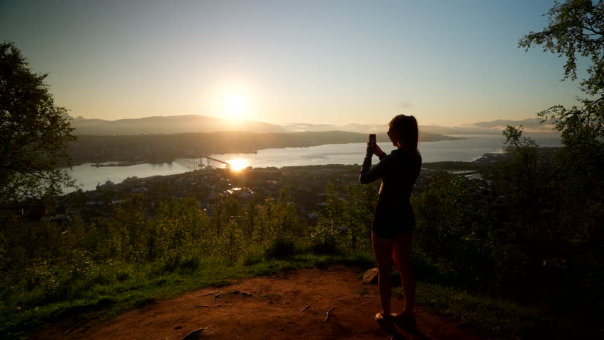 Female hiker captures sunset over Tromso at Fjellheisen lookout point on phone