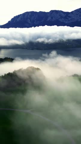 Vertical aerial parallax shot flying through morning fog and clouds revealing the beautiful Lake Toplitz and the Austrian Alps mountain range