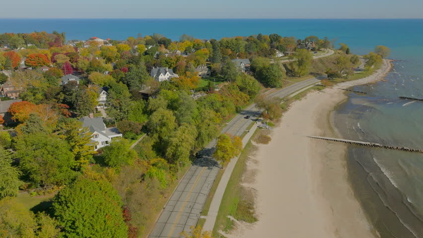 Drone aerial flying over shoreline road with cars driving past Sheboygan, Wisconsin neighborhood homes and fall trees beside Lake Michigan under a clear blue sky.