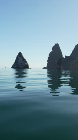 Sea, rocks, serene calm blue water reflecting ancient sea stacks in a peaceful ocean landscape under clear sky. Vertical video