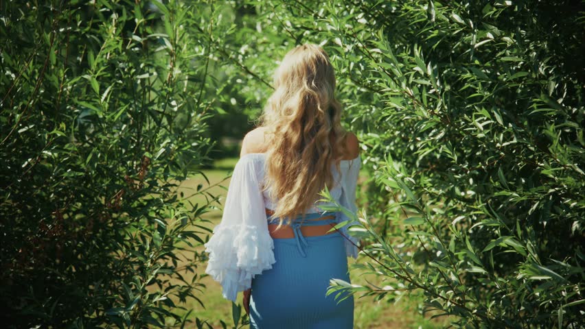 A woman with long hair walks through lush green foliage in a vibrant park. She wears a stylish top and skirt, enjoying the warmth of summer. The playful sunlight adds to the serene atmosphere.