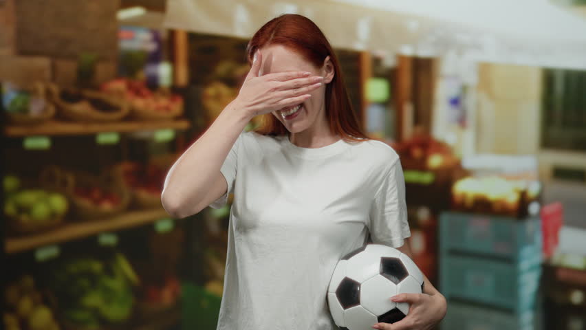 Woman with red hair smiling and holding a soccer ball in an outdoor market near fruit displays, covering her eyes playfully with her hand.