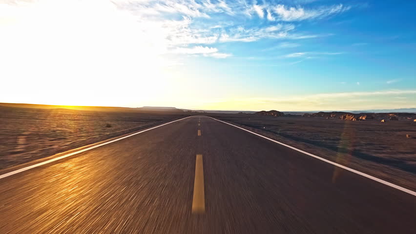 Driving car on asphalt road stretching through a vast desert landscape towards the horizon at sunset