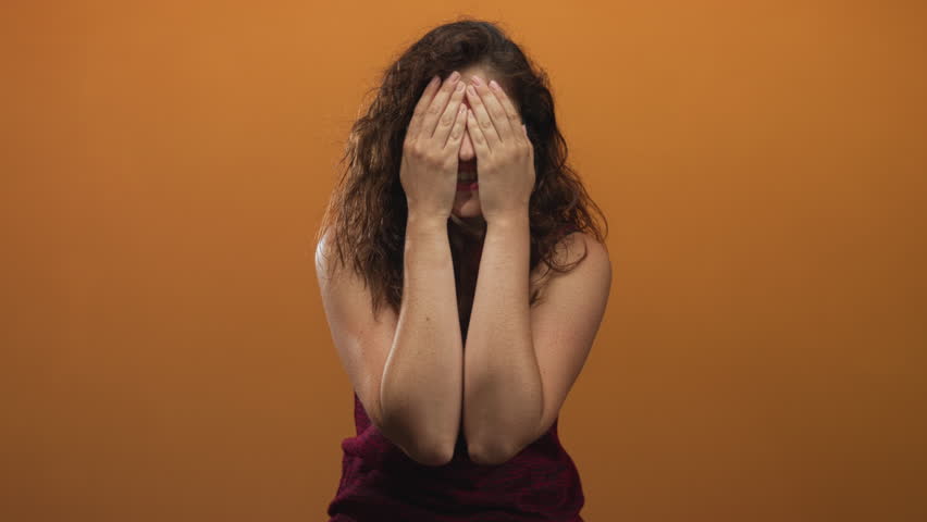 Young hispanic woman hands covering face in orange studio with mouth open and wide eyes; surprise reaction.