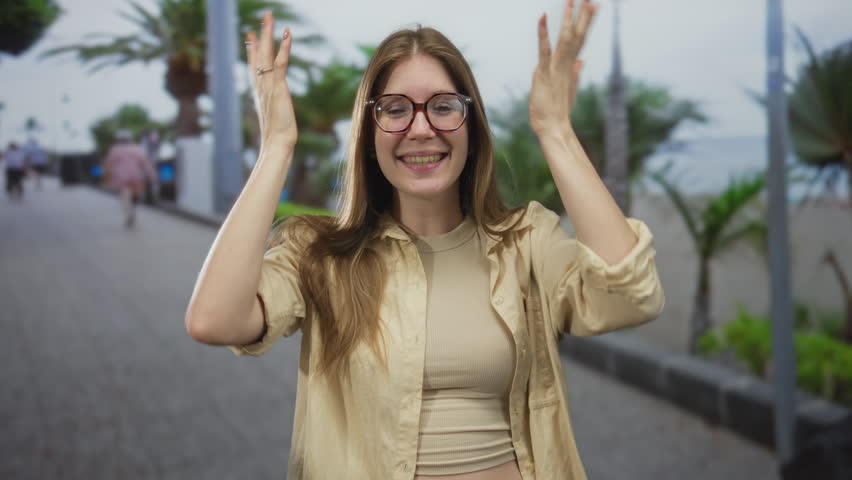 Woman in beige top with glasses places both hands on head while smiling near palm tree on street; inner joy.
