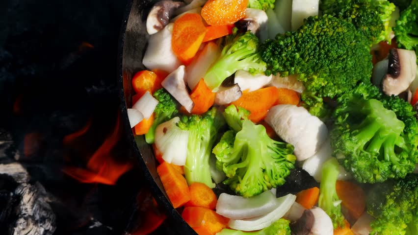 Detailed close-up of a black charred wok with vegetables simmering over a weak fire, with visible steam rising gently.