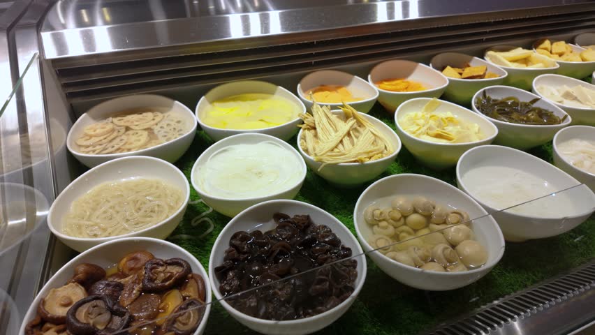 A buffet style food counter displaying various ingredients arranged in white bowls. The selection includes mushrooms, lotus root slices, noodles, tofu, and etc typically found in steamboat restaurant.