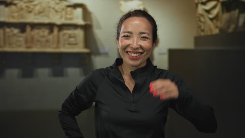 Woman smiling with hand to forehead, face visible in a museum building beside stone relief sculptures while wearing a black pullover and red nails; joy.