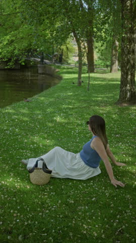 Woman relaxing in green park wearing sunglasses with handbag on lush grass under trees enjoying sunny day in peaceful garden setting.