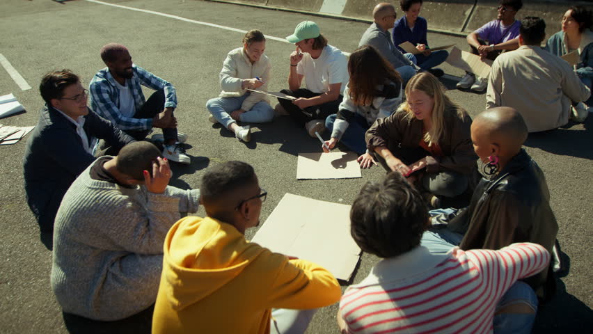 A dedicated youth group convenes outdoors, sitting in a circle on the asphalt to plan their next steps for a social cause. They are collaborating, discussing ideas, and preparing signs.