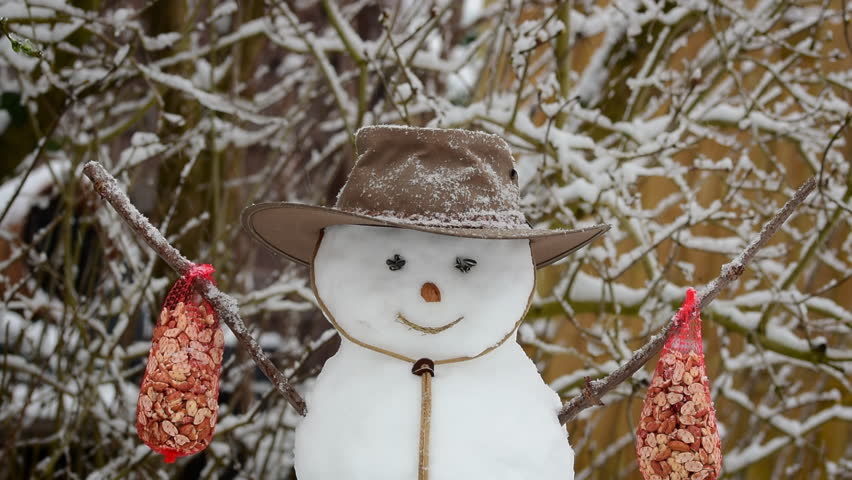 It snows on the smiling snowman wearing a hat and carrying peanuts food for the birds in winter