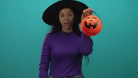 Woman pointing finger to halloween pumpkin bucket wearing witch hat and purple sweater in teal studio; playfulness. - Powered by Shutterstock - Get 15% off with code: PIKWIZARD15