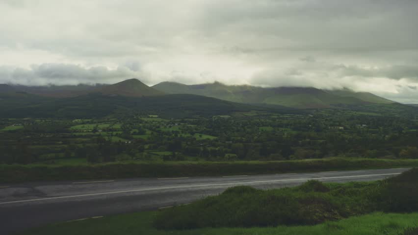 OA dramatic and moody landscape captures a panoramic view of rolling hills and mountains under a heavy, overcast sky. The dark, ominous clouds hang low, partially obscuring the mountain peaks and