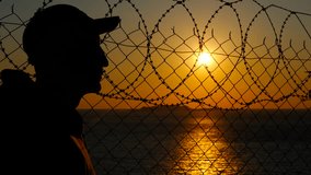 Man observing sunset behind barbed wire fence. Solitary male silhouette wearing cap, gazing across ocean horizon through barbed wire fence during golden sunset, symbolizing hope and freedom - Powered by Shutterstock - Get 15% off with code: PIKWIZARD15