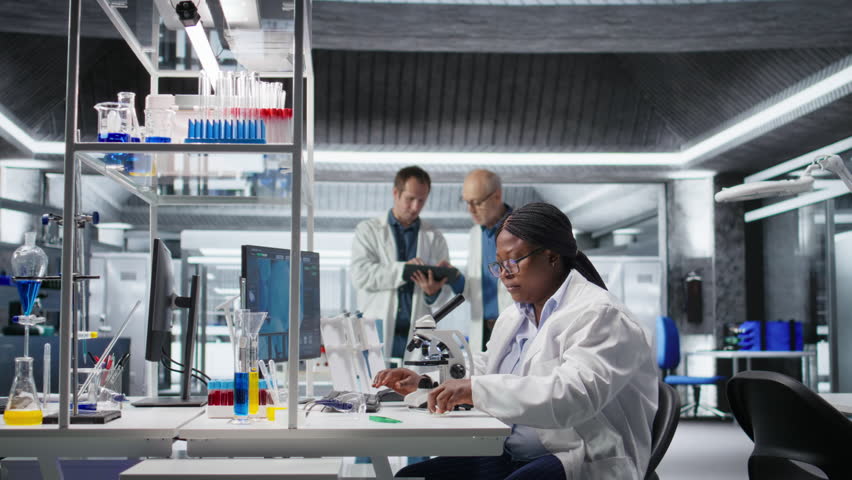 Woman working with microscope and sample tray in chemistry lab. Cellular investigation reflects the discovery process, medical research and nanotechnology for biotechnology solutions. Camera A.