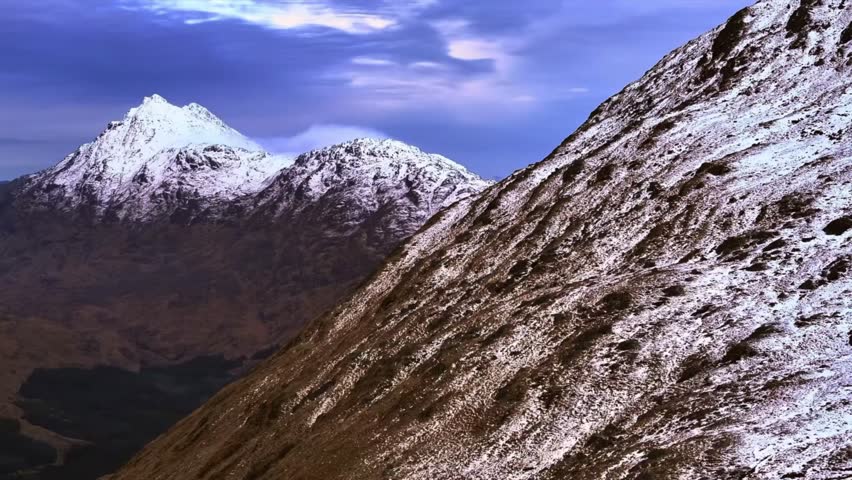 Rugged mountains dusted with snow rise sharply under a blue sky, showcasing steep slopes and jagged summits in a dramatic highland landscape.