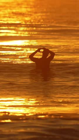 Slow motion vertical video of a male surfer sits on his surfboard and shows heart shape symbol, silhouetted by the sunset in Indian ocean.Maldives