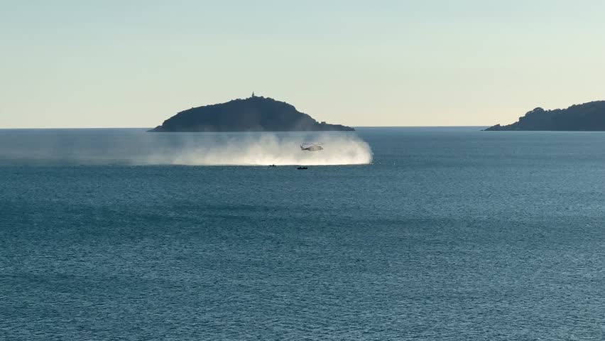 Helicopter hovering above the sea, creating dramatic spray near an island on the Ligurian coast in Italy. A seagull flies in the foreground, adding depth and motion to the scenic maritime landscape.