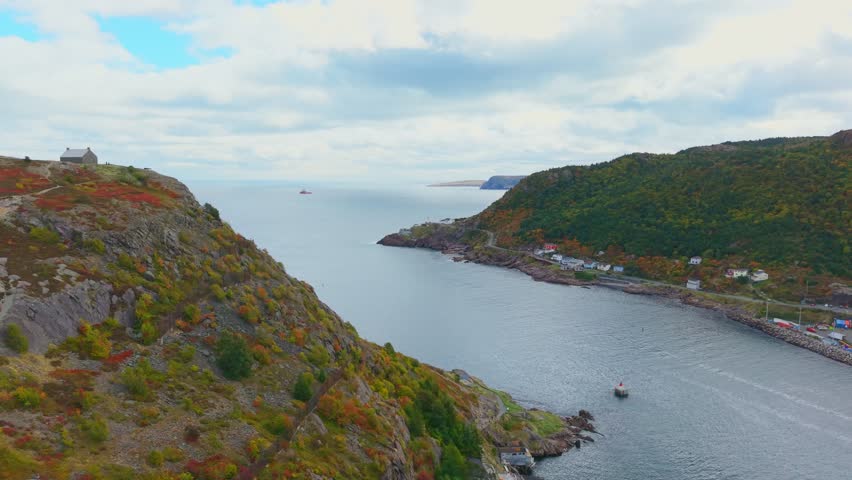 Wide aerial footage captures Fort Amherst’s serene bay nestled between rugged cliffs and forested hills, revealing calm turquoise waters, winding roadways and colourful houses in crisp autumn light.