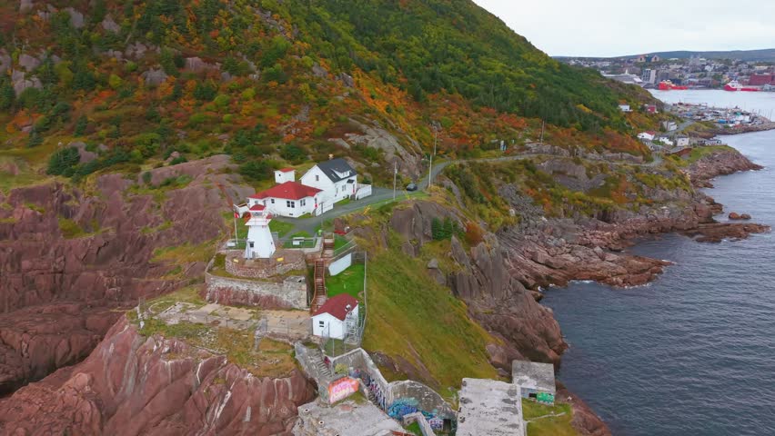 A sweeping drone view of Fort Amherst in Newfoundland during autumn captures the lighthouse, colorful houses, rugged cliffs and calm harbor under golden light.