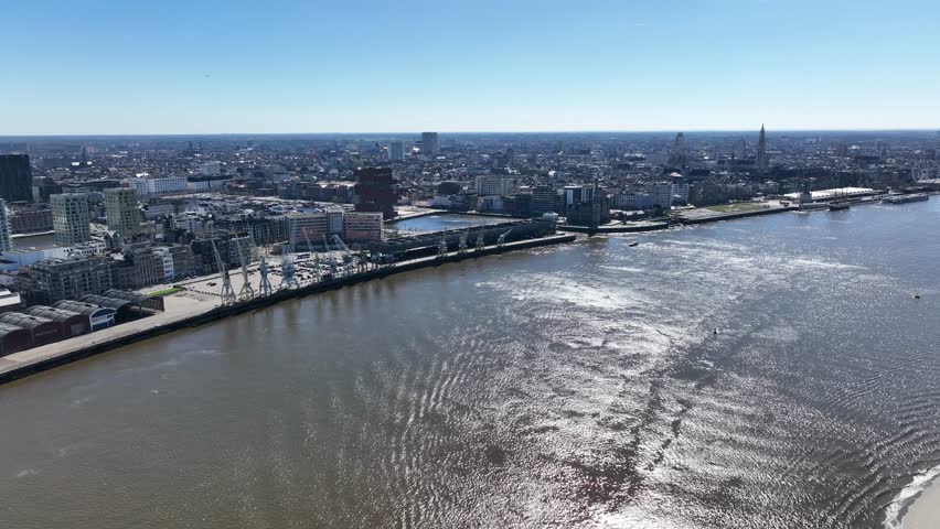 Pan left aerial shot of Antwerp skyline with Scheldt river, harbor docks, and cranes on a bright sunny day
