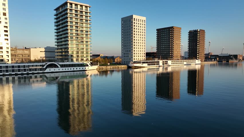 Zoom in on Antwerp modern residential towers reflecting in calm canal water with moored boats during golden hour light