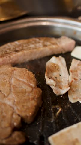 Tongs place garlic slice onto sizzling pork belly on tabletop Korean barbecue grill, warm lighting