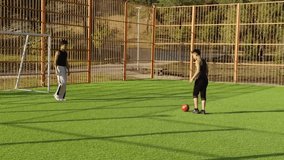 Two men on football pitch prepare for penalty. One lines up shot, goalkeeper observes with alert stance. Bright sunlight, sportswear, orange fence, red ball. Scene of strategy and rivalry. - Powered by Shutterstock - Get 15% off with code: PIKWIZARD15