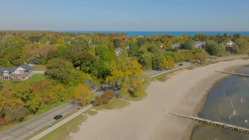 Drone aerial pushing over Lake Michigan beach toward a shoreline road with cars, neighborhood houses, and colorful autumn trees under a clear blue sky on a beautiful fall day.