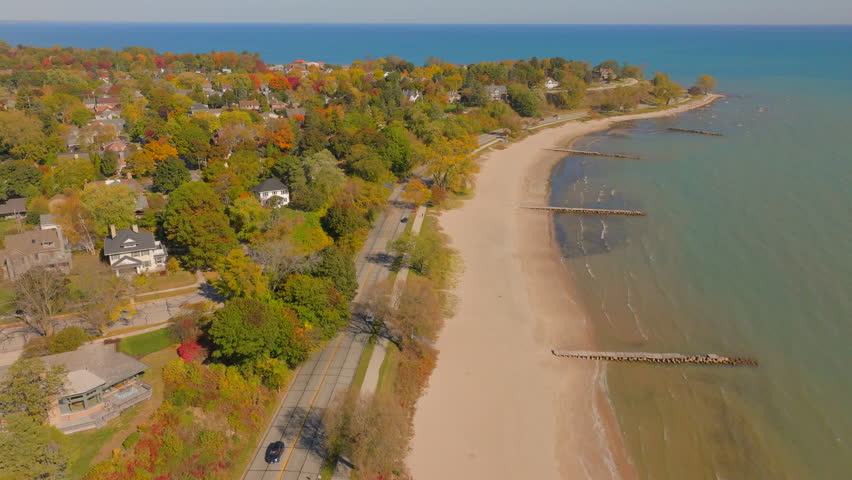 Drone aerial with pan and drift left over Lake Michigan beach toward Sheboygan, Wisconsin neighborhood homes and colorful autumn trees at peak foliage on a clear, pretty day.