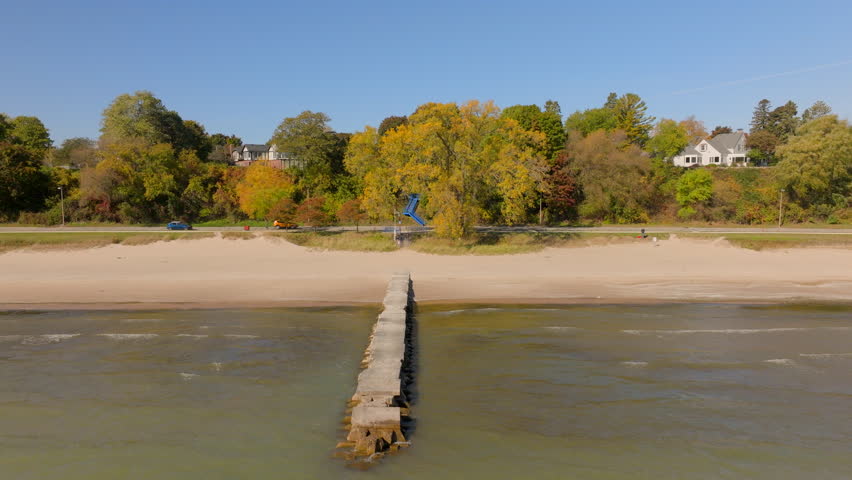 Drone aerial trucking left over Lake Michigan, showing beach, concrete pier, shoreline road, and Sheboygan neighborhood houses with peak fall color trees under a clear blue sky.