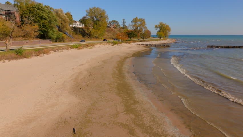 Drone aerial slow push over Lake Michigan beach with gentle waves and concrete pier toward shoreline road and Sheboygan neighborhood houses and autumn trees on a clear day.