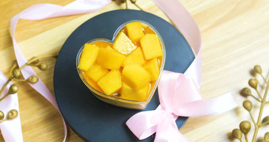 Heart shaped mango dessert sprinkled with golden accents, tied with pink ribbon, resting on dark plate against wooden background, symbolizing romantic sweetness