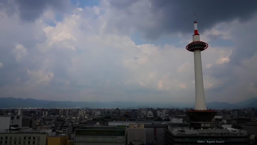 From Kyoto Station Skyway, the towering Kyoto Tower stands under rainy clouds, contrasting nicely with distant mountains and city buildings.