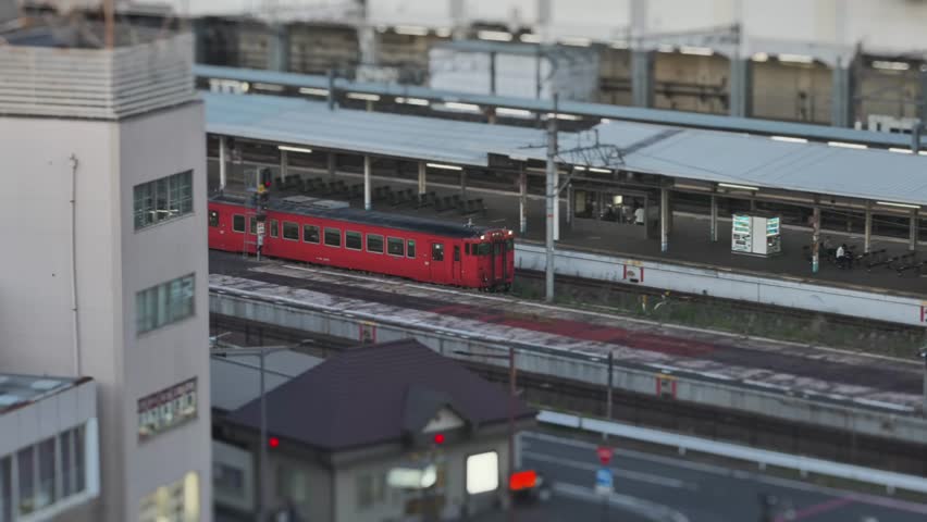 Overlooking Okayama Station, Japan: An old red train slowly enters a somewhat shabby platform with few passengers. The station and nearby buildings look calm under soft light.