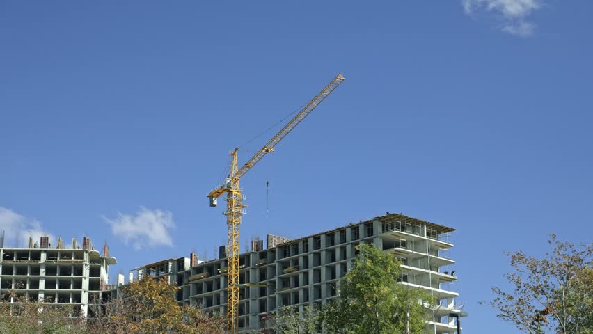 Yellow tower crane moving building materials on a large construction site against a clear blue sky. New residential complex under construction with concrete frame and workers on the upper floors