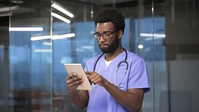 Confident african american doctor in blue coat using digital tablet standing in a modern hospital office. Focused medical worker physician in glasses chats online with patient or works in application - Powered by Shutterstock - Get 15% off with code: PIKWIZARD15